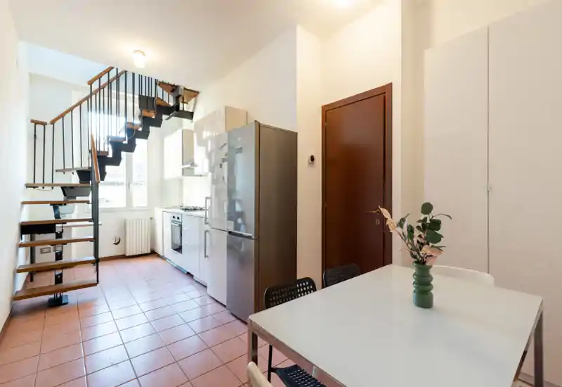 Cocina moderna y luminosa con electrodomésticos integrados, frigorífico independiente y mesa de comedor junto a la escalera — líneas limpias y luz natural que hacen el espacio funcional.