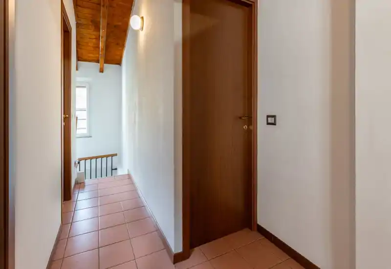 Pasillo estrecho y luminoso con suelo de terrazo rojo, paredes blancas y puertas de madera, que conduce a una escalera y una ventana con luz natural.