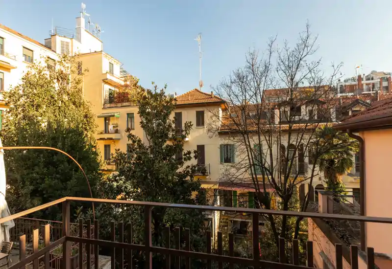 Balcón privado soleado con vistas a árboles maduros y edificios residenciales clásicos; ideal para el café de la mañana con luz natural cálida.