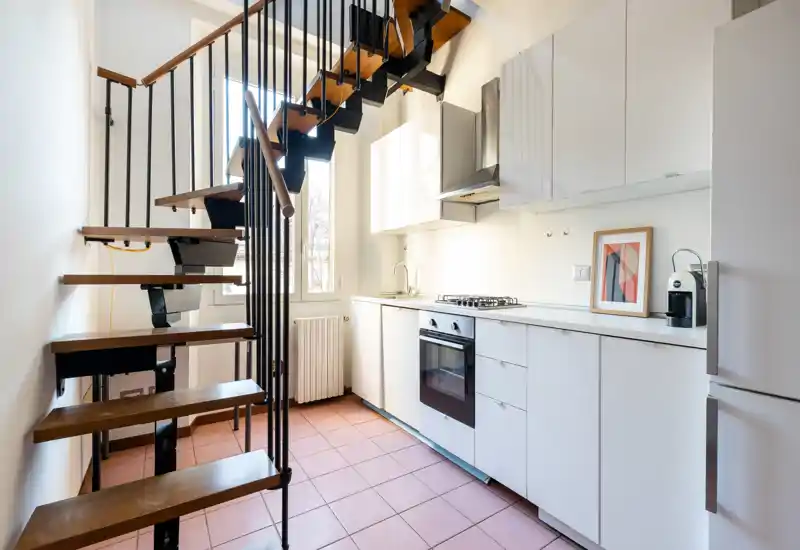 Bright modern kitchen with white cabinetry, built-in oven and gas hob, a compact sink and a coffee machine; an open wooden staircase runs along the left wall adding architectural interest.