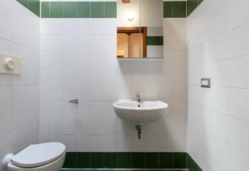Baño compacto y limpio con lavabo mural, armario con espejo e inodoro, azulejos blancos con franja verde.