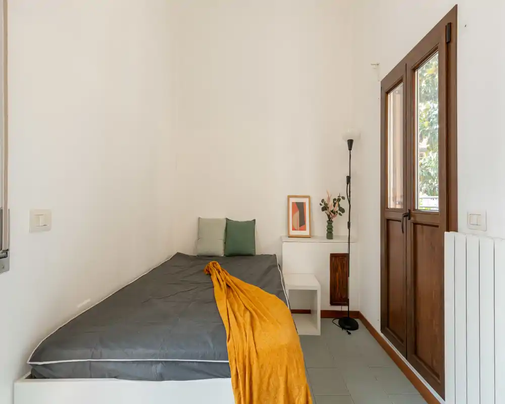 Compact, bright bedroom with a single bed dressed in grey linens and a mustard throw, small bedside table and tall window/door bringing natural light.