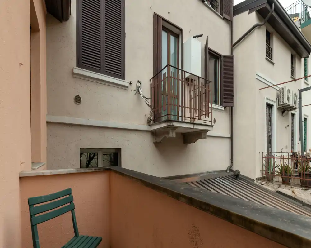 Small urban balcony view showing a peach-colored balustrade, a green folding chair and neighboring building facades with shutters and small balconies.