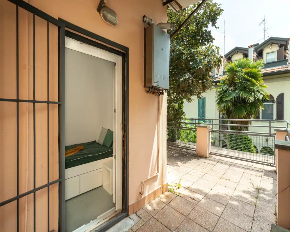 Sunny terrace with tiled floor and metal railing, looking out over neighboring houses and greenery; a small bedroom doorway opens onto the terrace.