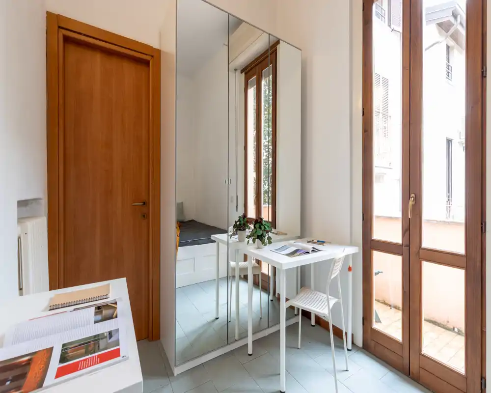 Small bright bedroom corner with a white desk, mirrored wardrobe and tall French windows that open to a balcony. The space feels modern and airy with natural light and minimal decorative touches.