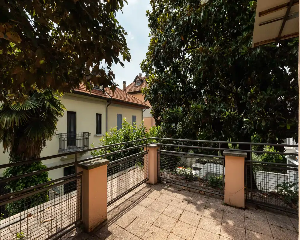 Sunny private terrace with tiled floor, metal railing and mature trees providing privacy — ideal for morning coffee or small outdoor seating.