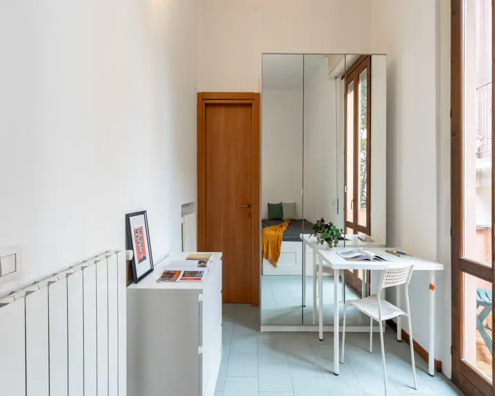 Compact, bright bedroom corner with a mirrored wardrobe reflecting the bed; a small desk and chair by a large window create a practical workspace. Neutral tones, simple furnishings and a warm throw add a welcoming touch.