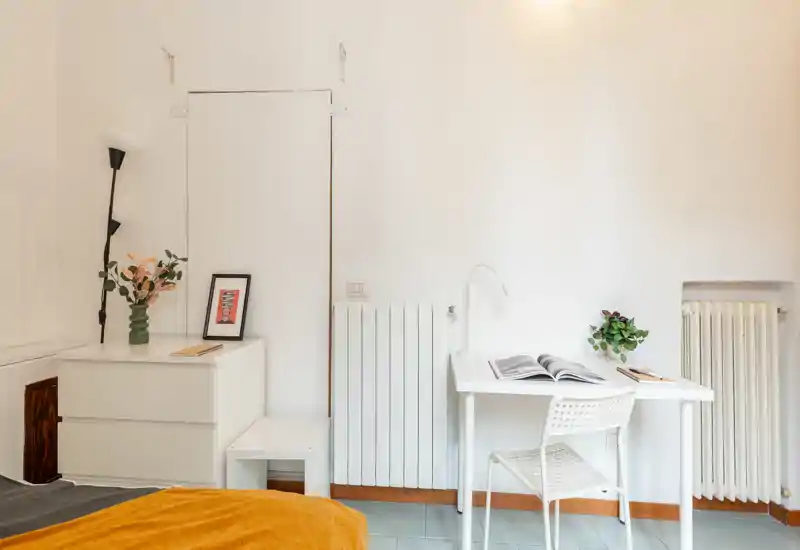 Coin chambre lumineux et minimaliste avec bureau blanc, chevet et lit visible avec couverture chaude — parfait pour une personne ou un étudiant.