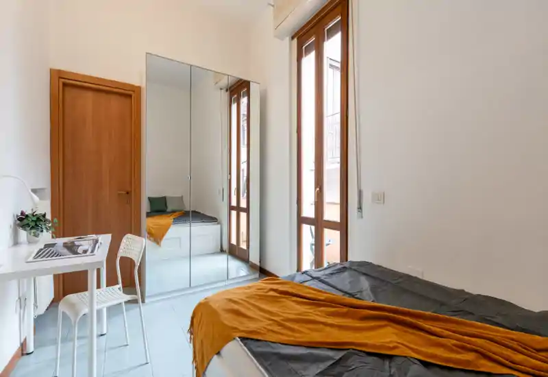 Chambre moderne et lumineuse avec lit double en linge foncé et plaid orange, armoire miroir et grandes fenêtres laissant entrer la lumière naturelle.