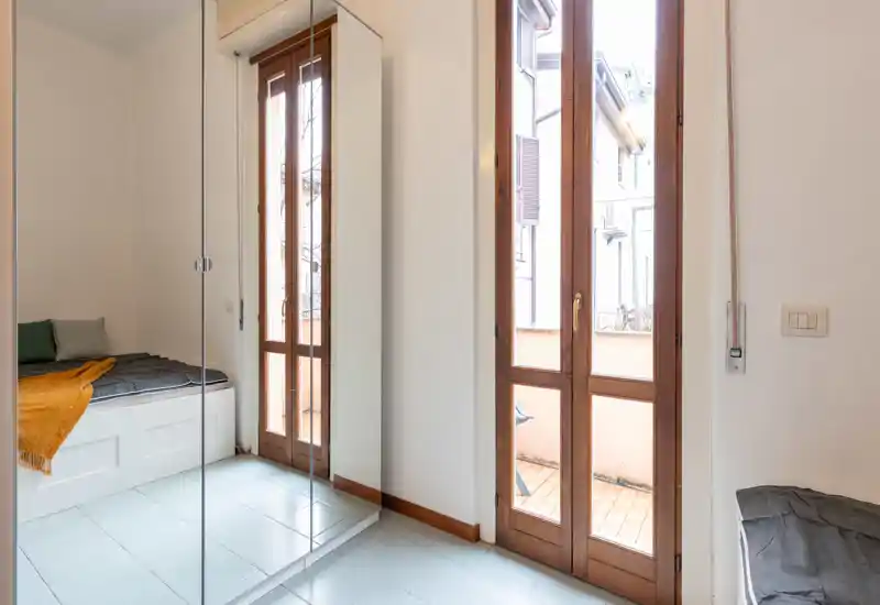 Bright, minimal bedroom with a built-in single bed, light tiled floor and full-height wooden glass doors opening to a small balcony. Crisp, neutral styling with a throw and cushions for comfort.