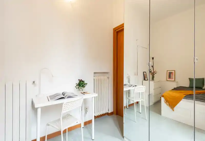 Chambre lumineuse et minimaliste avec lit blanc, plaid orange et placard miroir qui agrandit visuellement l’espace; un petit bureau offre un coin fonctionnel.