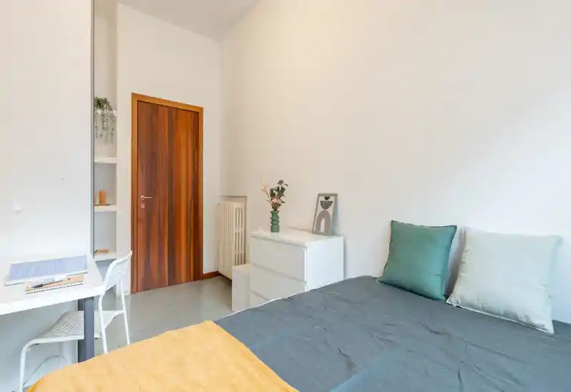 Chambre lumineuse et minimaliste avec un lit double bien fait, coussins verts et une petite commode blanche ; parfaite pour une personne seule ou un couple à la recherche d’un espace moderne.