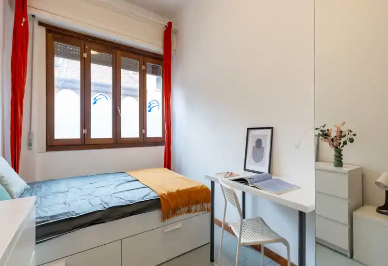 Chambre lumineuse et compacte avec un lit simple, des tiroirs de rangement intégrés, un petit bureau et une grande fenêtre apportant la lumière naturelle. Décor minimaliste avec une couverture moutarde et des rideaux rouges pour une touche de couleur.