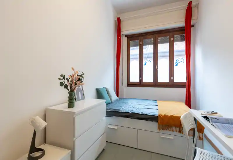 Chambre compacte et lumineuse avec lit simple surélevé doté de tiroirs de rangement, bureau près de la fenêtre et mobilier moderne et épuré. La grande fenêtre en bois apporte une belle lumière naturelle et les textiles colorés réchauffent l’ambiance.