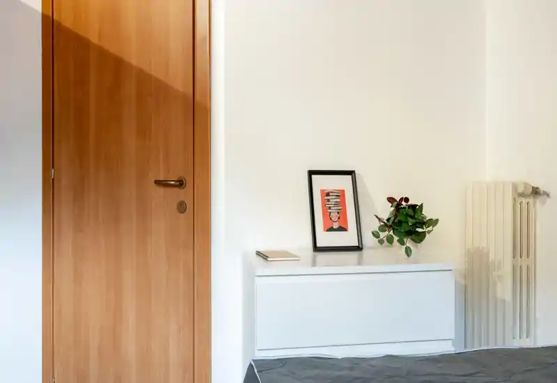 Minimal bedroom corner with a light wooden door, a low white chest, a radiator and a small visible portion of the bed; bright, clean and simply decorated with a framed print and a plant.