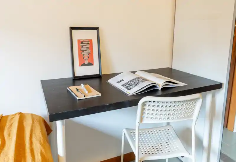 Small bedroom workspace with a dark wooden desk, open book, notebook and a white perforated chair next to a partially visible bed covered with a mustard throw.