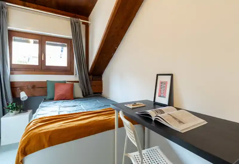 Chambre mansardée lumineuse avec un lit double compact, poutres en bois chaleureuses, table de chevet avec lampe et un long bureau idéal pour étudier ou travailler.