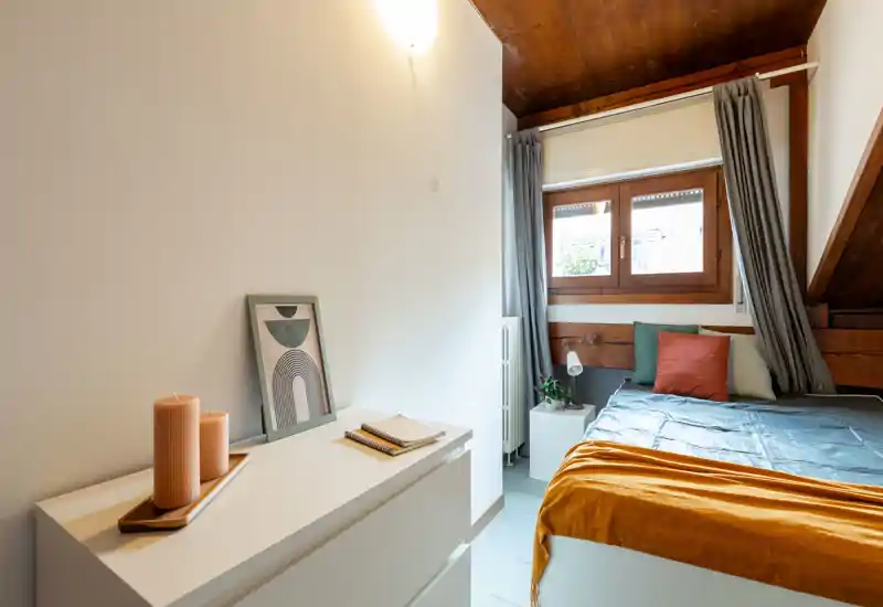 Chambre à coucher petite et chaleureuse avec lit bien fait, textiles colorés et lumière naturelle provenant d'une fenêtre en bois; décoration simple et commode blanche avec bougies pour une ambiance accueillante.