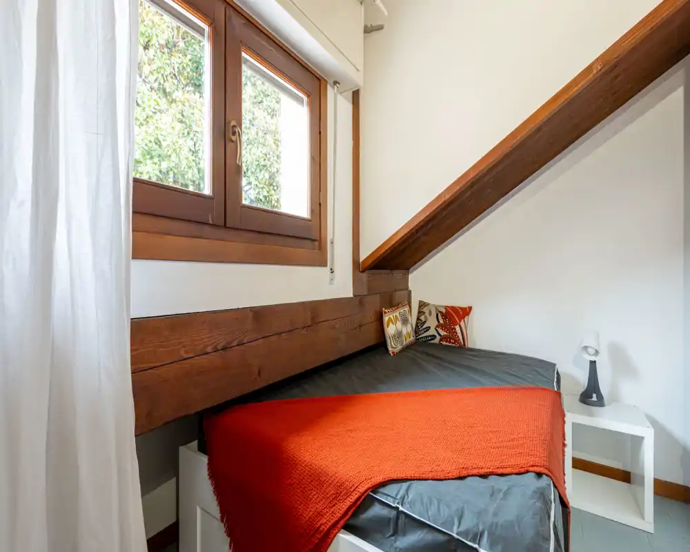 Chambre mansardée et cosy avec un lit simple sous une poutre en bois, textiles chaleureux et lumière naturelle par la fenêtre.