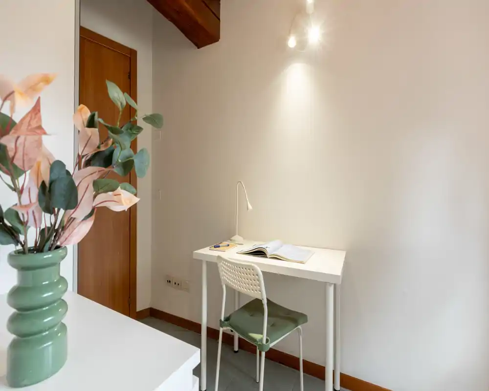 Coin bureau lumineux et minimaliste avec un bureau blanc, une chaise et une lampe de travail contre un mur neutre ; un vase décoratif et un coussin apportent une touche cosy.