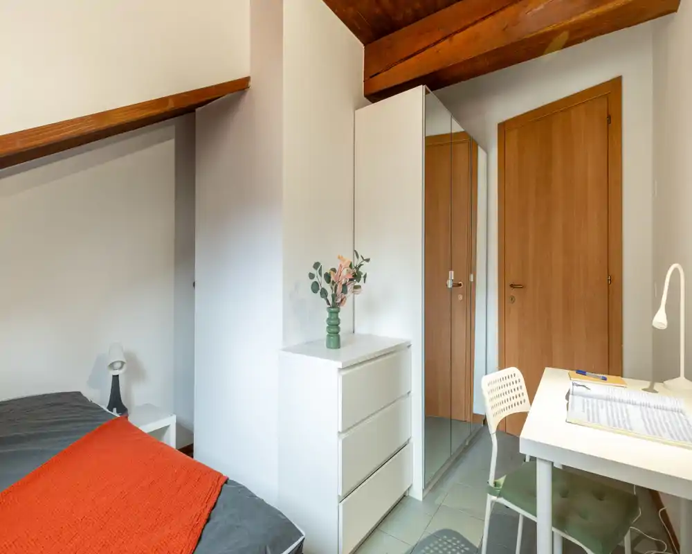 Chambre lumineuse et bien agencée avec lit visible, lampe de chevet, commode et petit bureau ; poutres en bois et armoire miroir apportent de la profondeur.