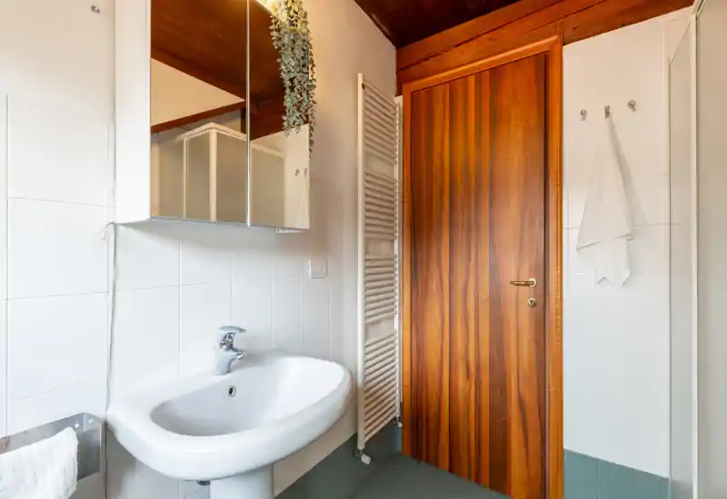 Bright, well-lit bathroom with a white sink, mirrored cabinet, towel radiator and wooden door — clean and functional space ideal for guests.