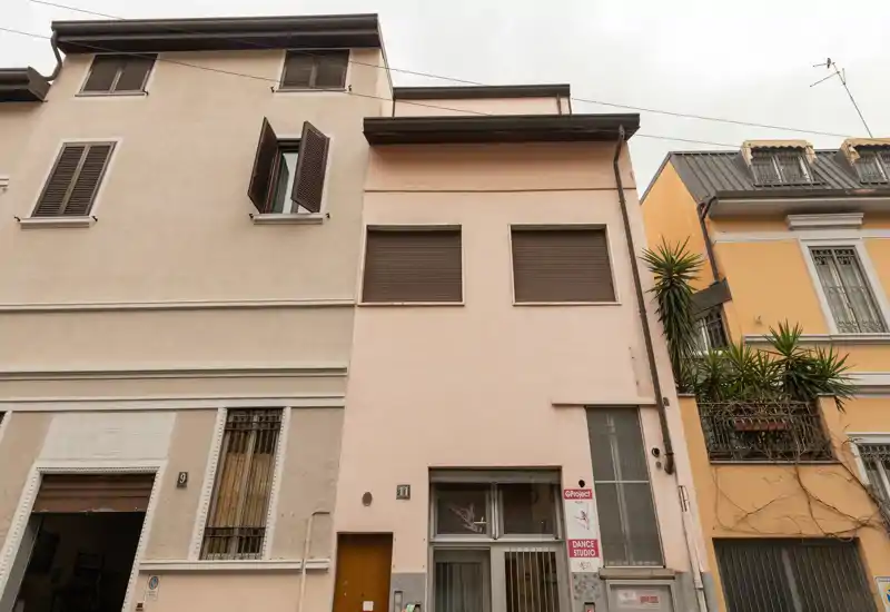 Exterior view of a narrow urban building facade with closed shutters and a ground-floor entrance, seen from the street under overcast light.