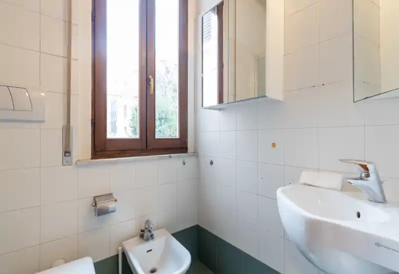 Small, bright bathroom with a sink, bidet and toilet visible; tiled walls, a wooden-framed window and a mirrored cabinet provide natural light and storage.