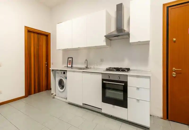 Compact modern kitchen with white cabinetry, built-in oven, gas hob, extractor hood and an integrated washing machine — practical and bright space ideal for a small apartment.
