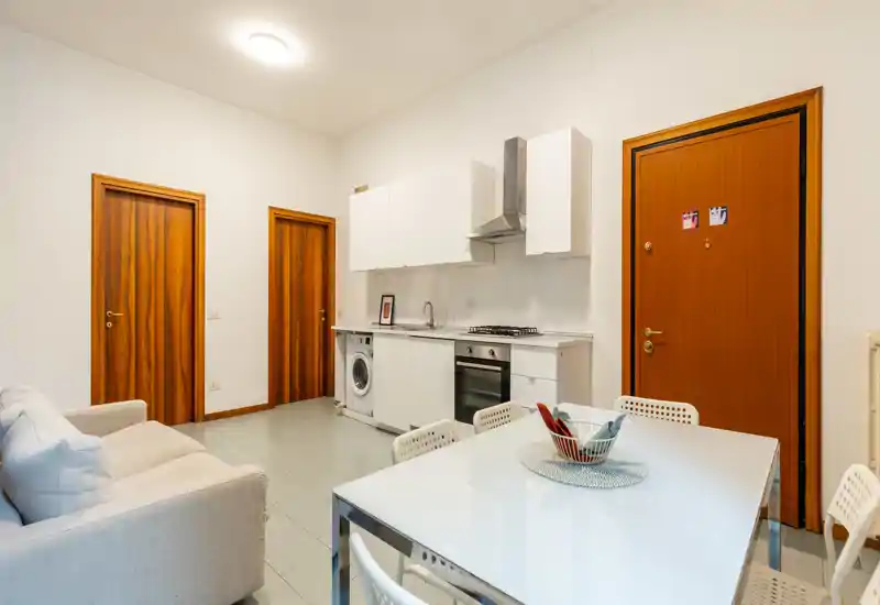 Bright open-plan living area with a compact white kitchenette, dining table and a small sofa — clean modern styling ideal for short rentals.