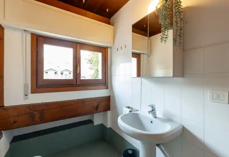 Small, bright bathroom with a pedestal sink, mirrored cabinet and a wooden-framed window providing natural light.