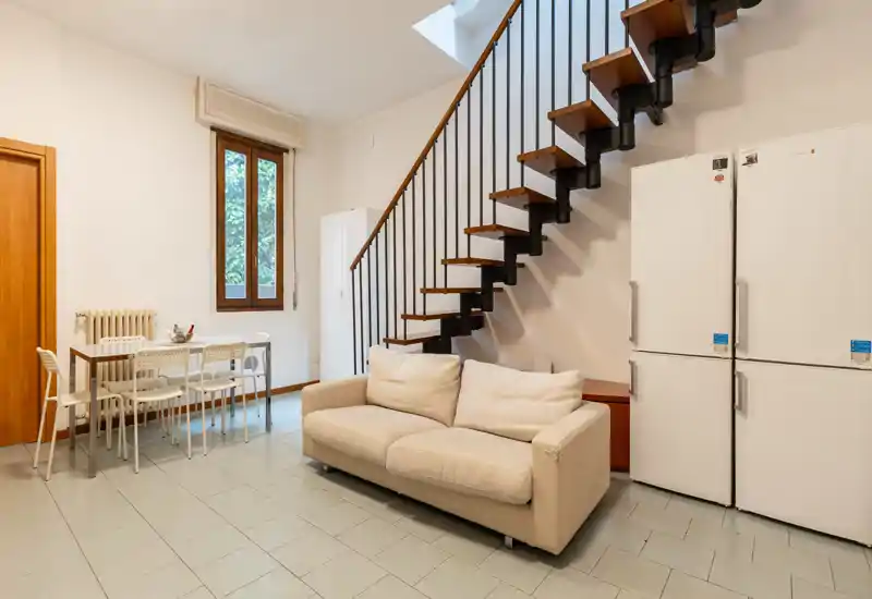 Bright, modern living area with a comfortable two-seater sofa, dining table and distinctive floating staircase — airy and well-lit, suitable for relaxed living.