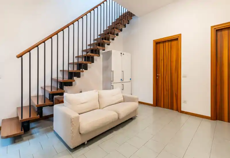 A compact living area featuring a light fabric sofa placed under a modern wooden staircase; warm wooden doors and neutral tiles create a clean, contemporary look.