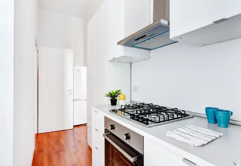Bright, modern galley kitchen with a gas cooktop, built-in oven and white cabinets. Clean styling, wooden floor and small decorative touches (plant, mugs) create a fresh, functional space.