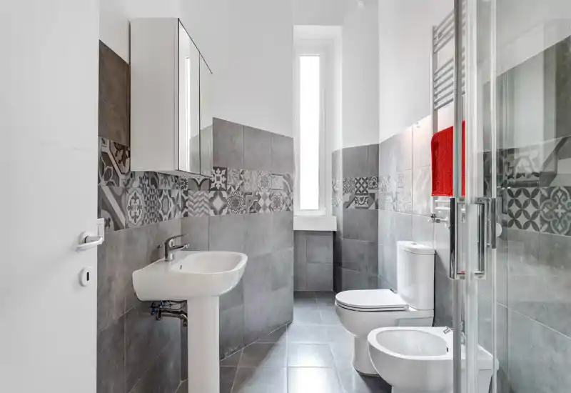 Modern, well-lit bathroom with grey patterned tiles, pedestal sink, toilet, bidet and a glass shower door; tall narrow window provides natural light.
