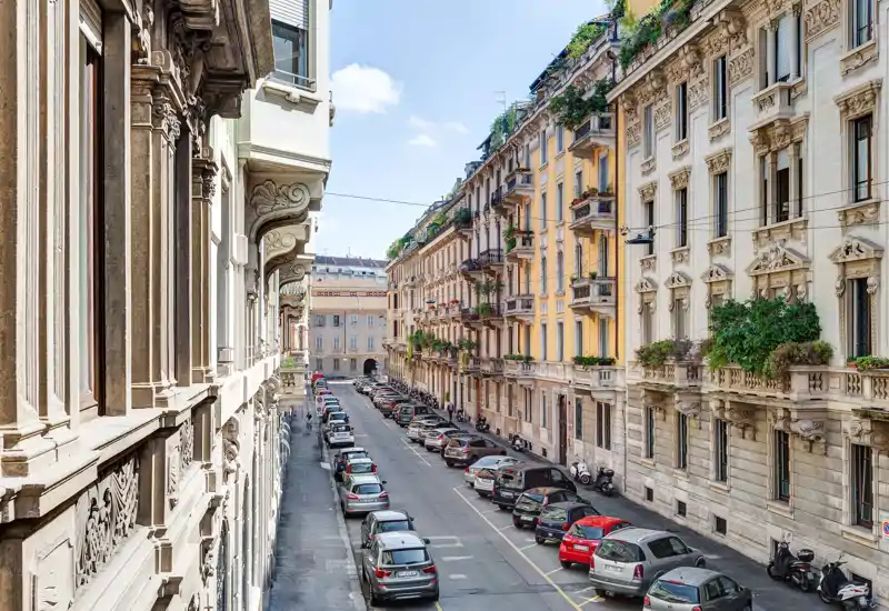 Sunny urban street view from a balcony showing classic ornate apartment façades, parked cars and a clear blue sky.