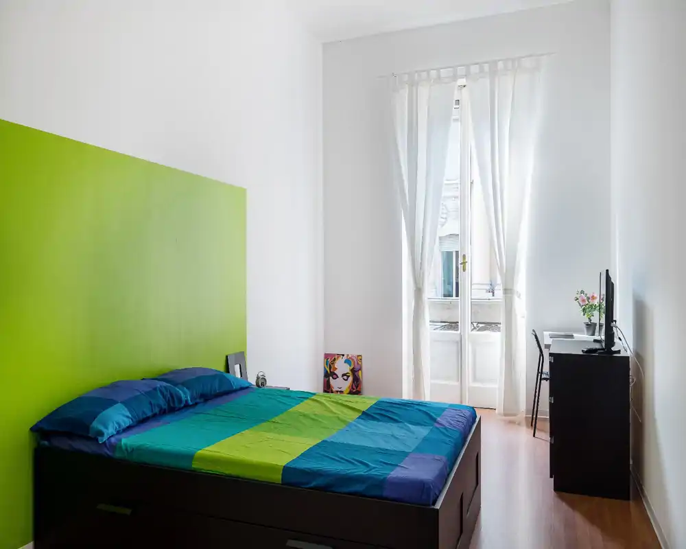 Dormitorio moderno y luminoso con una gran cama de ropa de cama colorida, ventana alta con cortinas traslúcidas y un pequeño escritorio con TV — limpio, bien iluminado y acogedor.