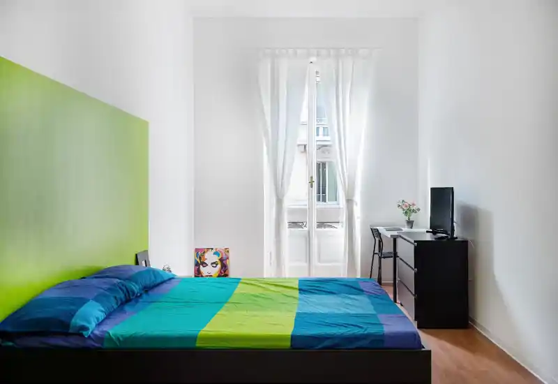 Bright, modern bedroom with a colorful double bed, tall sheer-window letting in abundant natural light, and a compact dresser with TV and small desk — ideal for a stylish single or couple.