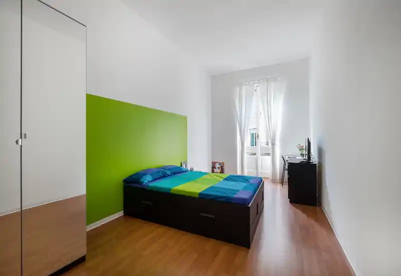 Bright, modern bedroom with a double bed featuring colorful linens, a green accent wall and natural light from a balcony window. Minimal, tidy furnishings include a wardrobe with mirrored door and a small desk with TV.