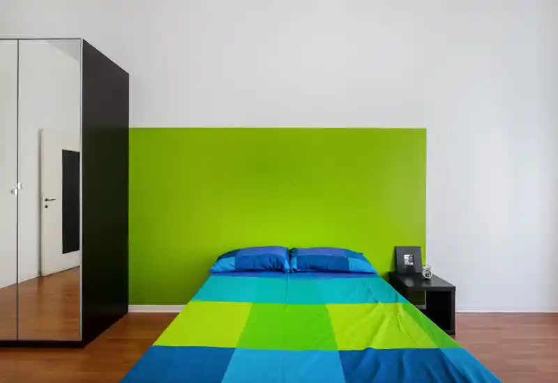 Bright, modern bedroom with a colorful blue-and-green bed against a vivid green accent wall, simple bedside table and mirrored wardrobe — clean minimalist styling ideal for listings.
