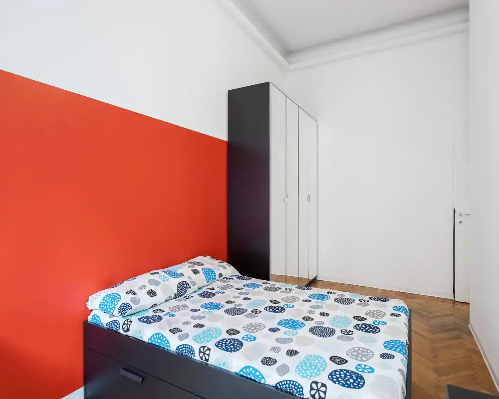 Camera da letto luminosa e moderna con letto matrimoniale dalla fantasia geometrica e una parete d'accento rossa. Armadio con ante a specchio e pavimento in parquet completano l'arredamento minimal.