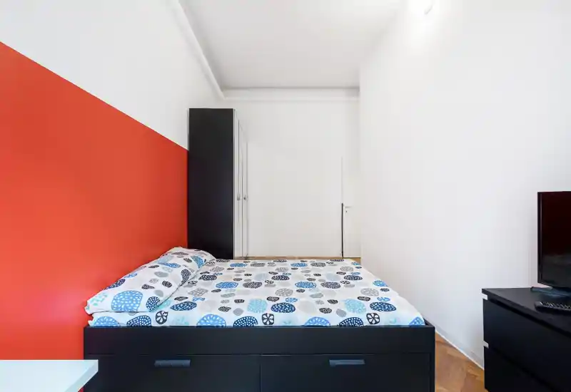 Bright, modern bedroom with a double bed, patterned bedding and a bold red accent wall. Simple black storage furniture and a clean, minimalist layout provide functional comfort.