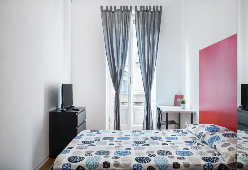 Bright, modern bedroom with a patterned double bed facing a tall window with sheer curtains; small desk and TV stand complete the minimalist layout.