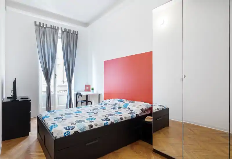 Bright modern bedroom with a double bed featuring patterned bedding, a bold red headboard panel, desk by the window and mirrored wardrobe — well lit and neatly arranged for rental photos.