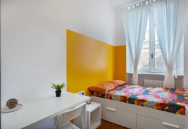 Bright, compact bedroom with a single bed against a yellow accent wall, colorful bedding, desk and window with sheer curtains letting in natural light.