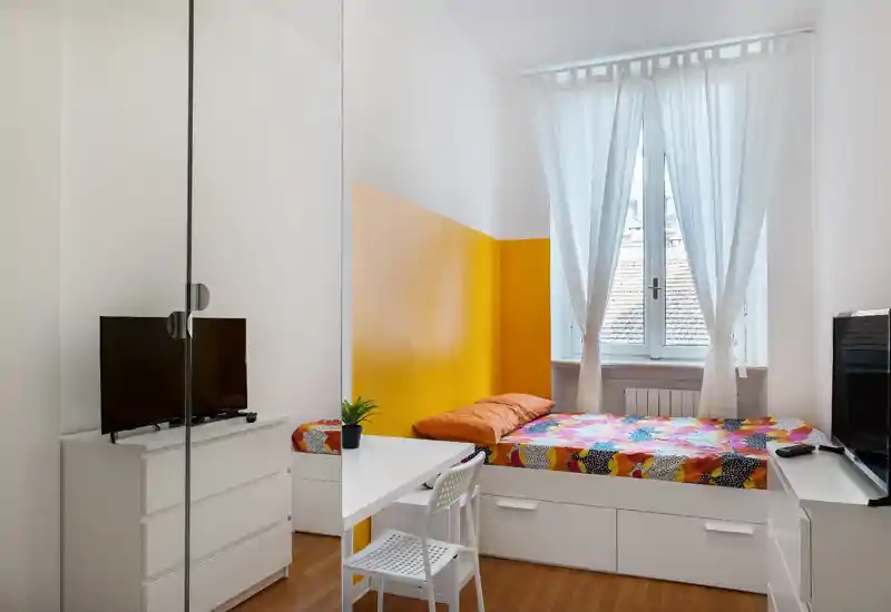 Bright single bedroom with a colorful bed, white storage drawers beneath the mattress, a small desk and TV — clean, modern and well lit by a large window.