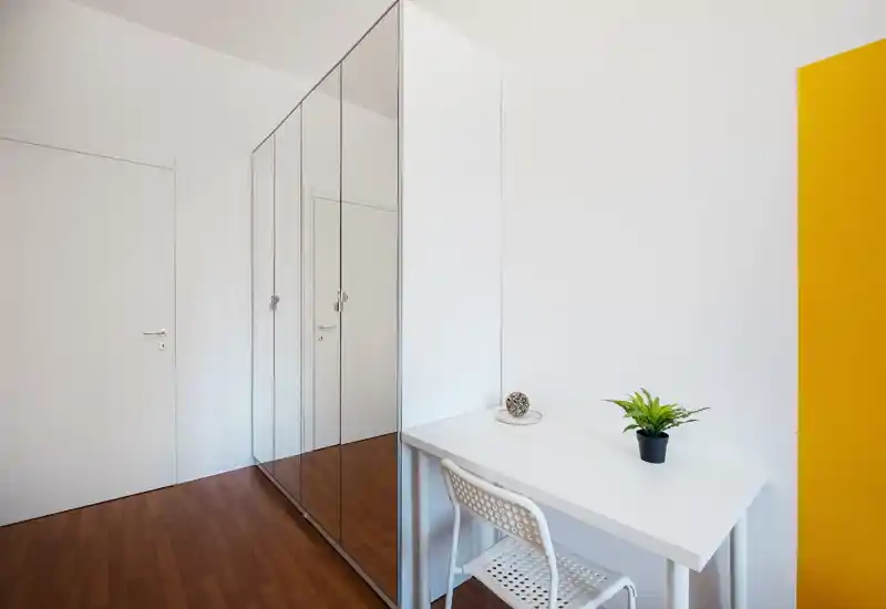 Minimal, bright room corner with a mirrored wardrobe and a small white desk with chair and plant; clean, modern styling ideal for a compact bedroom or study nook.