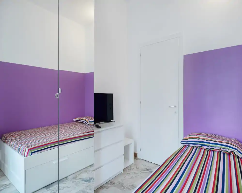 Bright, compact bedroom with two single beds, colorful striped bedding and a mirrored wardrobe that visually enlarges the space.