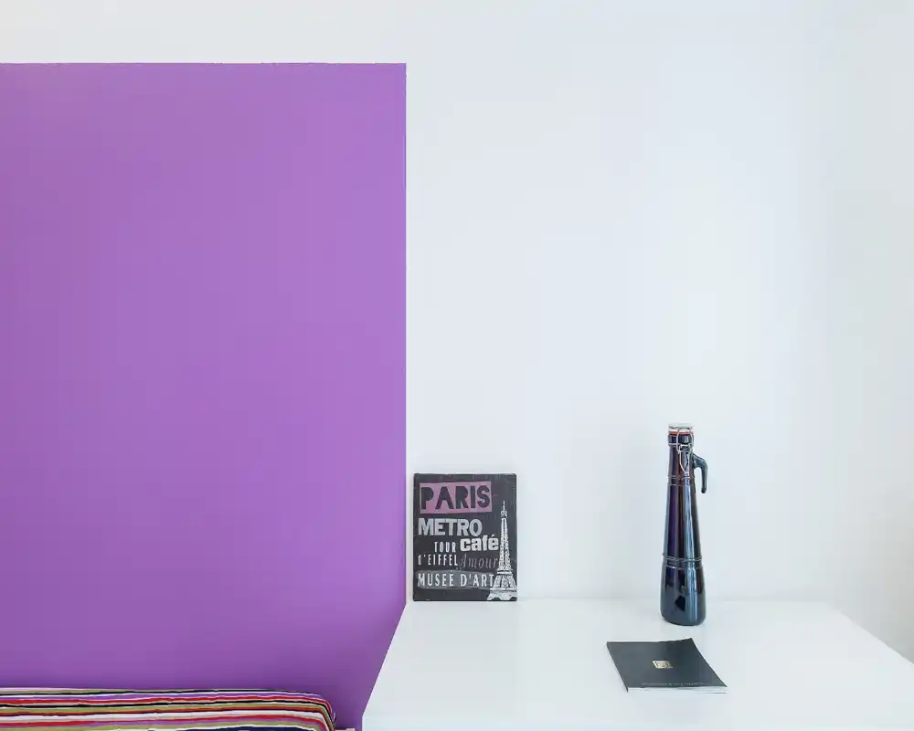 Minimal styled corner showing a purple accent wall, a low striped bed edge and a small desk with decorative items; bright, modern and neatly arranged.