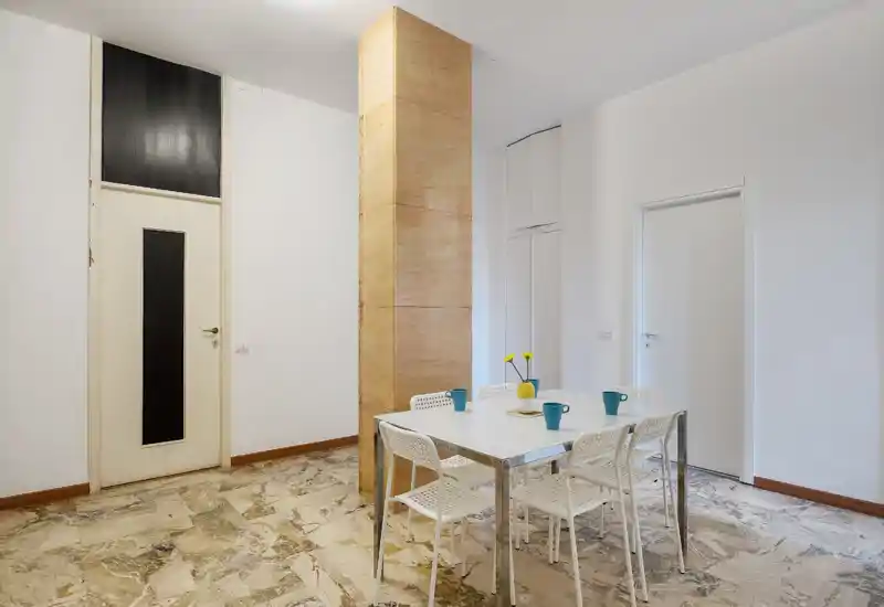 Simple, modern dining area with a white table and four chairs, bright walls, and a decorative vase with flowers on the table.